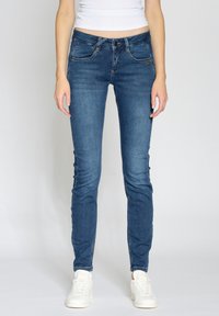 Přiléhavé modré džíny z denimu s pěti kapsami, zipem a jemným vyblednutím. V kombinaci s bílými sportovními teniskami.