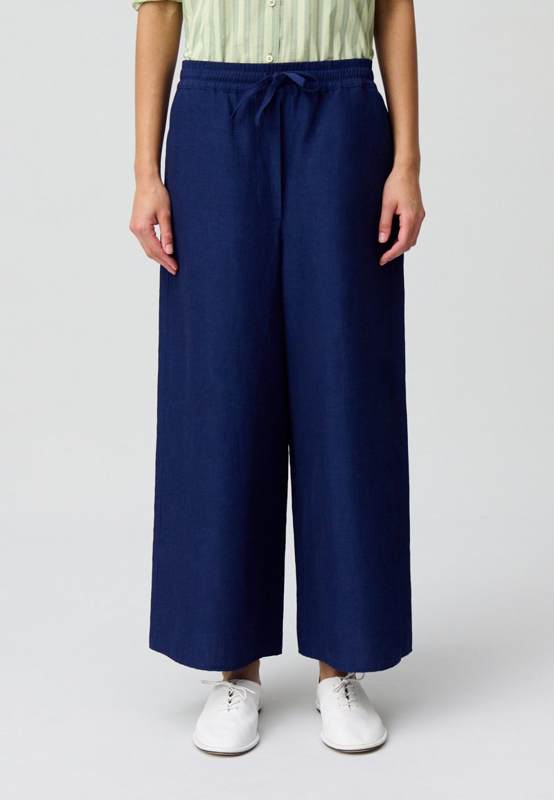 Pantaloni a gamba larga blu navy con vita con coulisse, realizzati in un tessuto leggero; abbinati a scarpe bianche, che evidenziano una vestibilità rilassata e uno stile informale.