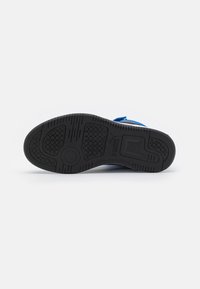 Semelle en caoutchouc noir d'une chaussure Puma avec des motifs de bande de roulement et un logo visible, inclinée horizontalement sur un fond blanc uni.