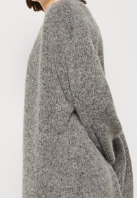 Personne portant un pull gris texturé en maille ample à manches longues, vue de profil sur un fond blanc uni.