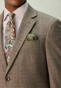 Brauner karierter Blazer mit Kerbkragen, ein gestreiftes rosa-weißes Hemd, gemusterte Krawatte in Blau, Rot und Grün sowie ein grünes Einstecktuch.