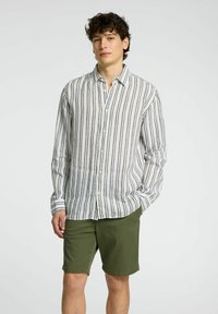 Camicia di lino a righe in bianco, blu navy e beige, con bottoni e maniche lunghe. Abbinata a pantaloni corti verde oliva, il modello sta in piedi con le mani in tasca.