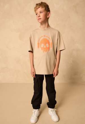 Niño rubio de pie, con camiseta beige con un gráfico de calavera naranja y auriculares, pantalones cargo negros y zapatillas blancas, fondo neutro.