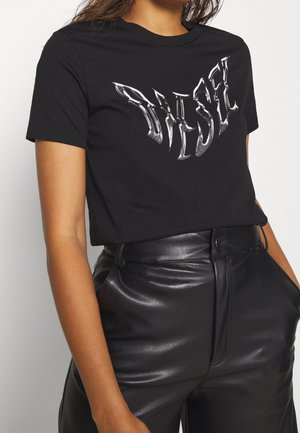 Kobieta ubrana w czarny t-shirt z krótkim rękawem z zniekształconym białym napisem "DIESEL" oraz czarne skórzane spodnie z wysokim stanem, z dłońmi w kieszeniach.