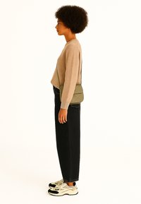 Beige gebreide trui, hoge zwarte jeans, olijfgroene schoudertas en veelkleurige sneakers, gezien vanaf de zijkant.