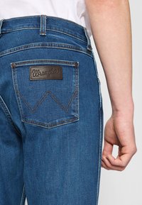 Jeans de mezclilla en azul oscuro con un parche de cuero con el logo de Wrangler en el bolsillo trasero. Presenta costuras en contraste y un diseño clásico de cinco bolsillos.