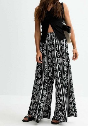 Frau trägt ein schwarzes ärmelloses Oberteil und weite schwarze Hosen mit weißen Blumenstreifen, hält eine schwarze und beige Schultertasche.