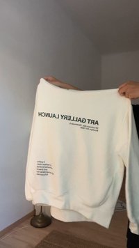 Fehér kereknyakú pulóver, fekete nyomtatott szöveggel a hátsó oldalon, amely egy művészeti galéria megnyitóját részletezi, bordázott mandzsettával és alsó szegéllyel.