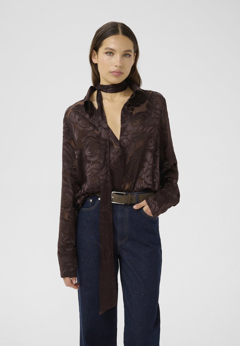 Camicia a maniche lunghe, marrone scuro, con un motivo floreale. Presenta un colletto, un dettaglio con cravatta ed è abbinata a pantaloni in denim a vita alta.