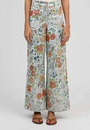 Femme portant un pantalon large à motif floral multicolore, associé à des sandales marron à brides sur un fond uni.