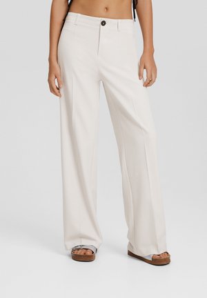 Pantalon blanc taille haute à jambes larges avec plis sur le devant, fermeture par bouton, porté avec des sandales argentées sur un fond neutre.