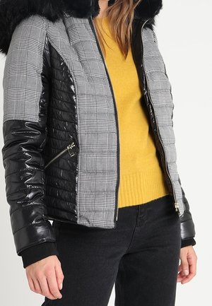 Kvinde iført en sort og grå ternet dunjakke med sorte ærmer og en pelskrave over en gul sweater og sorte bukser.