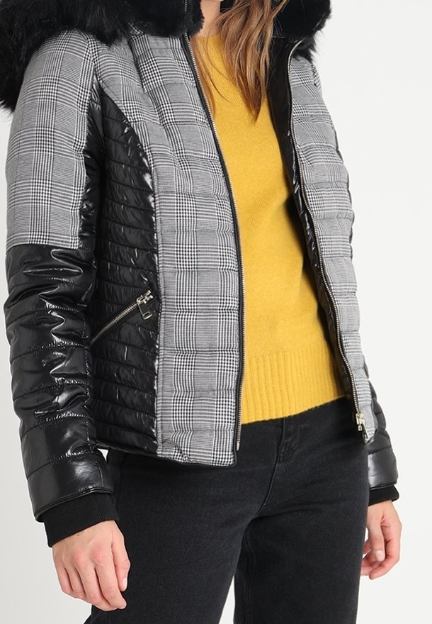 Femme portant une doudoune à carreaux noirs et gris avec des manches noires et un col en fourrure par-dessus un pull jaune et un pantalon noir.