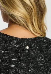 Svart och vit stickad tröja med ett strukturerat mönster, bred halsringning och en liten metallisk detalj på övre delen av ryggen.