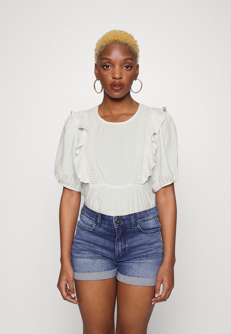 Blusa verde-clara com mangas com franja e frente texturizada, combinada com calções de ganga com bainha dobrada. Design simples, ajuste descontraído.