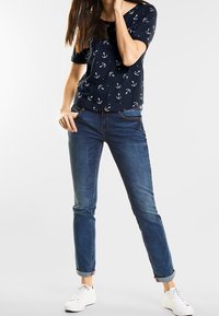 Marinblå t-shirt med vitt ankermönster, kombinerad med ljusblå jeans, lätt uppvikta ärmar och vita sneakers. Avslappnad outfit.