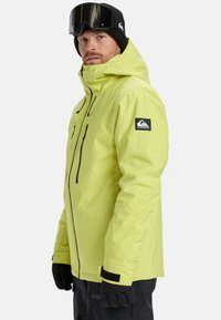 Heldergeel waterdichte skijas met zwarte ritsen en accenten, voorzien van een hoge kraag, zakken en een logo-patch op de mouw.