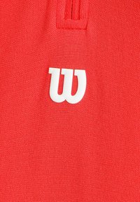 Camisa polo de punto roja con una textura de punto y un logotipo 'W' estilizado en color blanco en la zona del pecho.