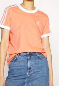 Coral short-sleeve t-shirt with white ribbed neckline and cuffs. Features three white stripes on shoulders and an embroidered logo on chest.