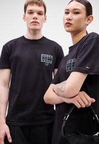 Czarne bawełniane koszulki z przodu z naszywką z logo, okrągłym dekoltem i luźnym krojem. Jeden model trzyma czarną torebkę.