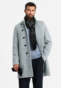 Abrigo de mezcla de lana de color gris claro con seis botones negros, cuello a medida y bolsillos laterales. Combinado con una bufanda de cuadros negros y guantes.