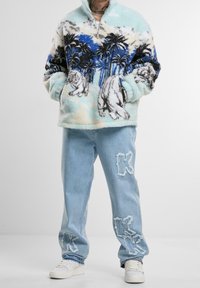 Fuzzy, light blue quarter-zip pullover with palm tree and bear print; paired with distressed, light denim jeans featuring frayed "K" accents.