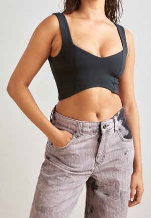 Kobieta w czarnym krótki topie na ramiączkach i wysokim stanem szarymi jeansami z efektem sprania, z rękami w kieszeniach na neutralnym tle.