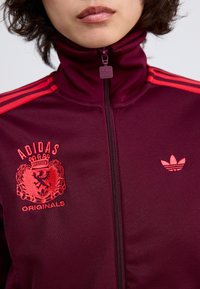 Vínová zipová bunda Adidas s červenými pruhy, obsahující velký znak a malé vyšívané logo. Hladká textura a těsně přiléhající design.