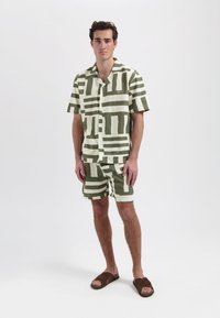 Knoop-up kortarm shirt en bijpassende short in crème en olijfgroen met abstracte blokpatronen; platte sandalen aan de voeten.