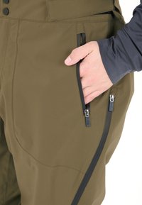 Olivenfarvede isolerede bukser har en glat tekstur med lynlåslommer, der er fremhævet af sorte metaldele, og de er designet til funktionalitet.