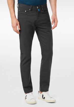 Mann trägt dunkelgraue Slim-Fit-Hose und weiße Sneakers mit schwarzen und beigen Details, steht vor schlichtem hellem Hintergrund.