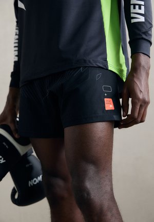 Schwarze Sportshorts mit geometrischem Design und orangefarbenem "Venum"-Label. Kombiniert mit einem langärmligen schwarzen und grünen Oberteil, während er Boxhandschuhe hält.
