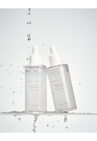 Due bottiglie di vetro traslucido con contagocce bianchi, etichettate "NEEDLY Ampoule", contenenti un liquido chiaro, che schizzano nell'acqua.