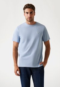 Hombre vestido con una camiseta de manga corta azul claro con un pequeño logo en forma de triángulo en el pecho, jeans azul oscuro, de pie contra un fondo claro y sencillo.
