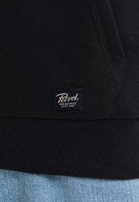 Maglione nero con polsini e fondo a coste. Presenta un tessuto testurizzato e un'etichetta blu con testo bianco, che recita "Petrol Industries Since 1989."