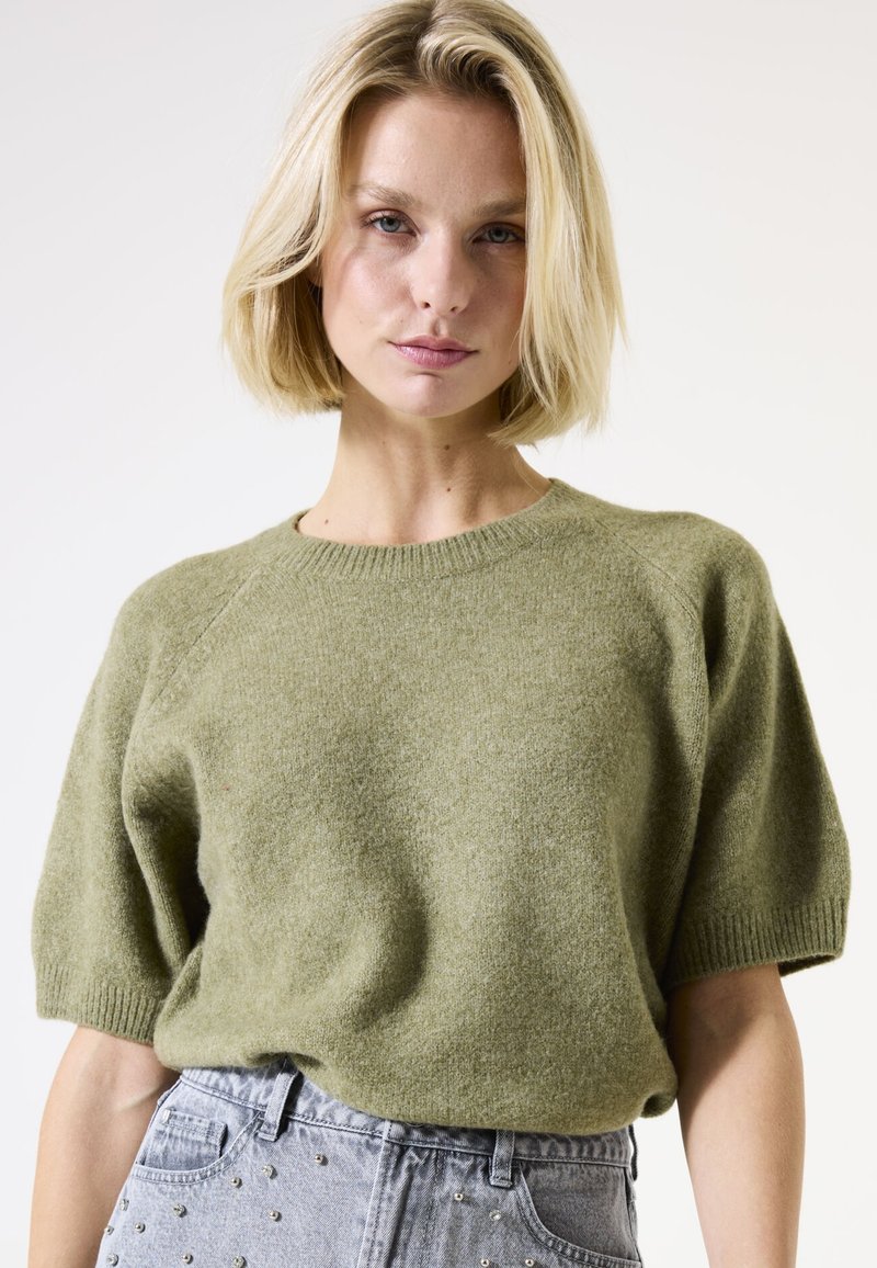 Pull en torsades vert olive avec des manches courtes et un col côtelé, associé à un jean taille haute bleu clair avec des clous en argent.