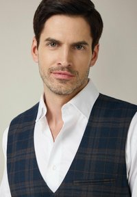 Granatowa kamizelka w kratę o gładkiej fakturze, z ostrym dekoltem i kieszenią, noszona na białej koszuli z klasycznym kołnierzykiem.