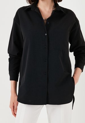 Woman wearing a loose black button-up shirt with chest pocket and silver chain necklace, styled with white pants, hands in pockets.