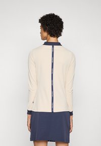 Light beige long-sleeve top with navy collar and hem. Features a central navy logo stripe along the back. Smooth fabric, comfortable fit.