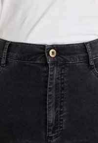 Zwarte corduroy broek heeft een fitted ontwerp, een gouden knop bij de taille en zichtbare textuur ribbels in de stof.