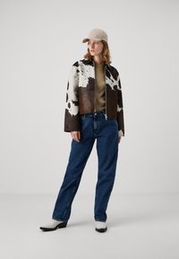 Braun-weiße Kuhfelljacke, olivgrünes Oberteil, blaue Jeans, beiger Hut und weiße Cowboystiefel mit schwarzen Sohlen. Schlichte, moderne Silhouette.