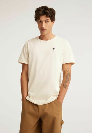 Młody mężczyzna z kręconymi włosami, ubrany w ecru t-shirt z małym logo byka i brązowe spodnie, stoi z rękami w kieszeniach.
