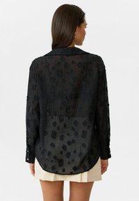 Vrouw in een zwarte doorschijnende blouse met lange mouwen en getextureerde vierkantpatronen, gecombineerd met een crèmekleurige geplooide minirok, met haar rug naar de camera gekeerd.