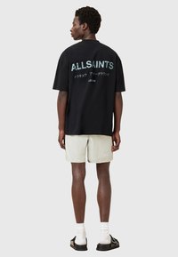 Black short-sleeve shirt with large white "ALLSAINTS" text on the back. Worn with light beige shorts and black footwear.