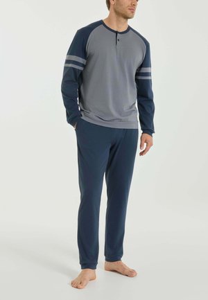 Uomo che indossa una maglia henley a maniche lunghe grigia e blu navy con maniche rigate e pantaloni blu navy, in piedi a piedi nudi su uno sfondo chiaro uniforme.