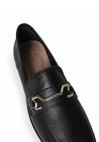 Black leather loafers with a smooth texture, featuring a metallic geometric accent on the strap. Subtle branding visible inside.