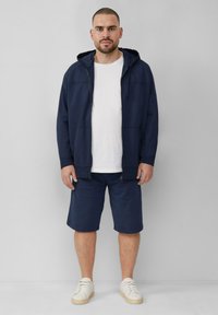 Marineblauwe zip-up hoodie met voorzakken, gecombineerd met bijpassende marineblauwe shorts. Daaronder een effen wit T-shirt en lichtgekleurde sneakers.