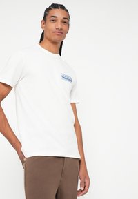 Biała koszulka z krótkim rękawem z bawełny, z niebieskim logo "RIP CURL" na lewej piersi. Połączona z brązowymi spodniami w luźnym kroju.
