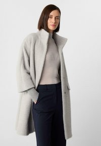 Donna in piedi con indosso un cappotto testurizzato grigio chiaro sopra un maglione a collo alto grigio e pantaloni navy scuri, con una mano in tasca.