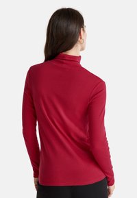 Frau mit langen dunklen Haaren, die ein enges rotes Rollkragenoberteil und schwarze Hosen trägt, von hinten vor einem weißen Hintergrund gezeigt.